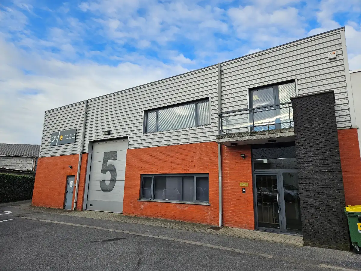 Een modern industrieel gebouw met een groot cijfer 5 op een grijze garagedeur, oranje bakstenen onderwanden en metalen bovenwanden. Er zijn grote ramen, een glazen ingang en buiten staat een gele afvalbak. De lucht is gedeeltelijk bewolkt.