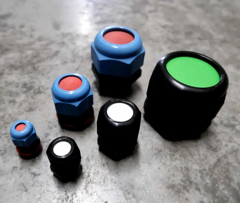 Zes drukknoppen van verschillende afmetingen en kleuren – rood, wit en groen – zijn aangebracht op een grijs oppervlak met structuur. De knoppen hebben een zwarte of blauwe behuizing.