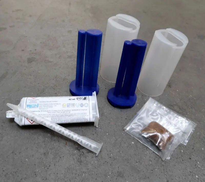Een tweecomponenten-epoxylijmset met een dubbele spuit, een mengmondstuk, twee plastic bakjes met blauwe deksels en een zakje met bruin materiaal, allemaal op een grijs oppervlak.