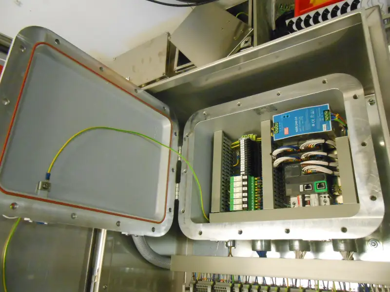Een open metalen schakelkast toont bedrading, connectoren, een printplaat en een blauw apparaat. Het deksel staat open, waardoor de interne onderdelen en een aardingsdraad die aan de kast is bevestigd, zichtbaar zijn.