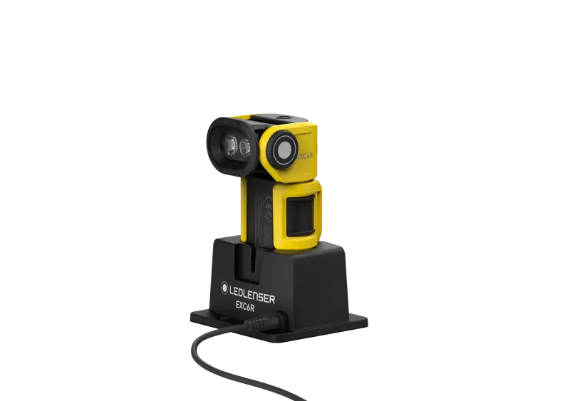 Een compacte geel-zwarte LEDLenser EX7R-zaklamp staat rechtop in zijn zwarte oplaadstation, met een kabel die in het oplaadstation is aangesloten, tegen een effen witte achtergrond.