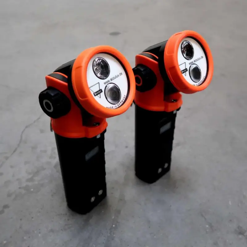 Twee draagbare noodzaklampen met een oranje en zwarte behuizing, schuin geplaatste lampkoppen en dubbele lenzen, die rechtop staan op een grijs betonnen oppervlak.