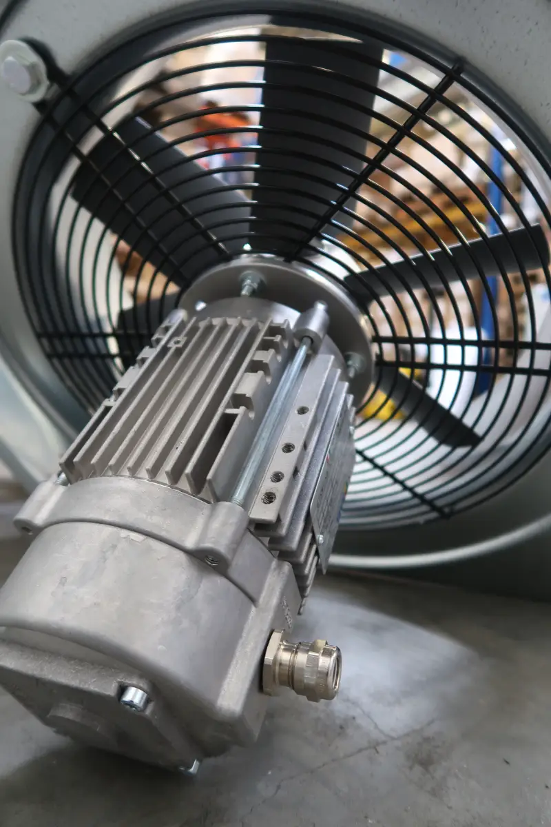 Close-up van een grote elektromotor voor een zwarte metalen ventilatorbeveiliging, waarbij de metalen behuizing en de onderdelen in detail te zien zijn; de motor staat op een betonnen ondergrond in een industriële omgeving.