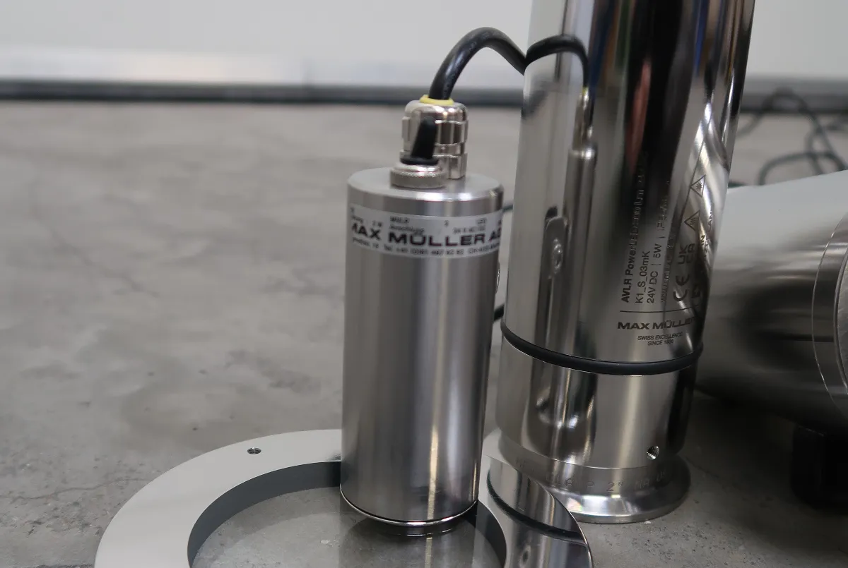 Een close-up van een cilindervormige industriële sensor van roestvrij staal met het opschrift MAX MÜLLER, die op een betonnen vloer naast andere metalen apparatuur staat.