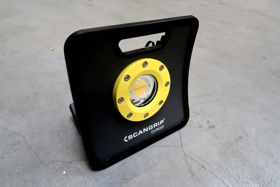 Op een grijze betonnen vloer staat een zwart-gele draagbare Scangrip-werklamp. De lamp heeft een vierkant frame met een handvat aan de bovenkant en een ronde LED in het midden.
