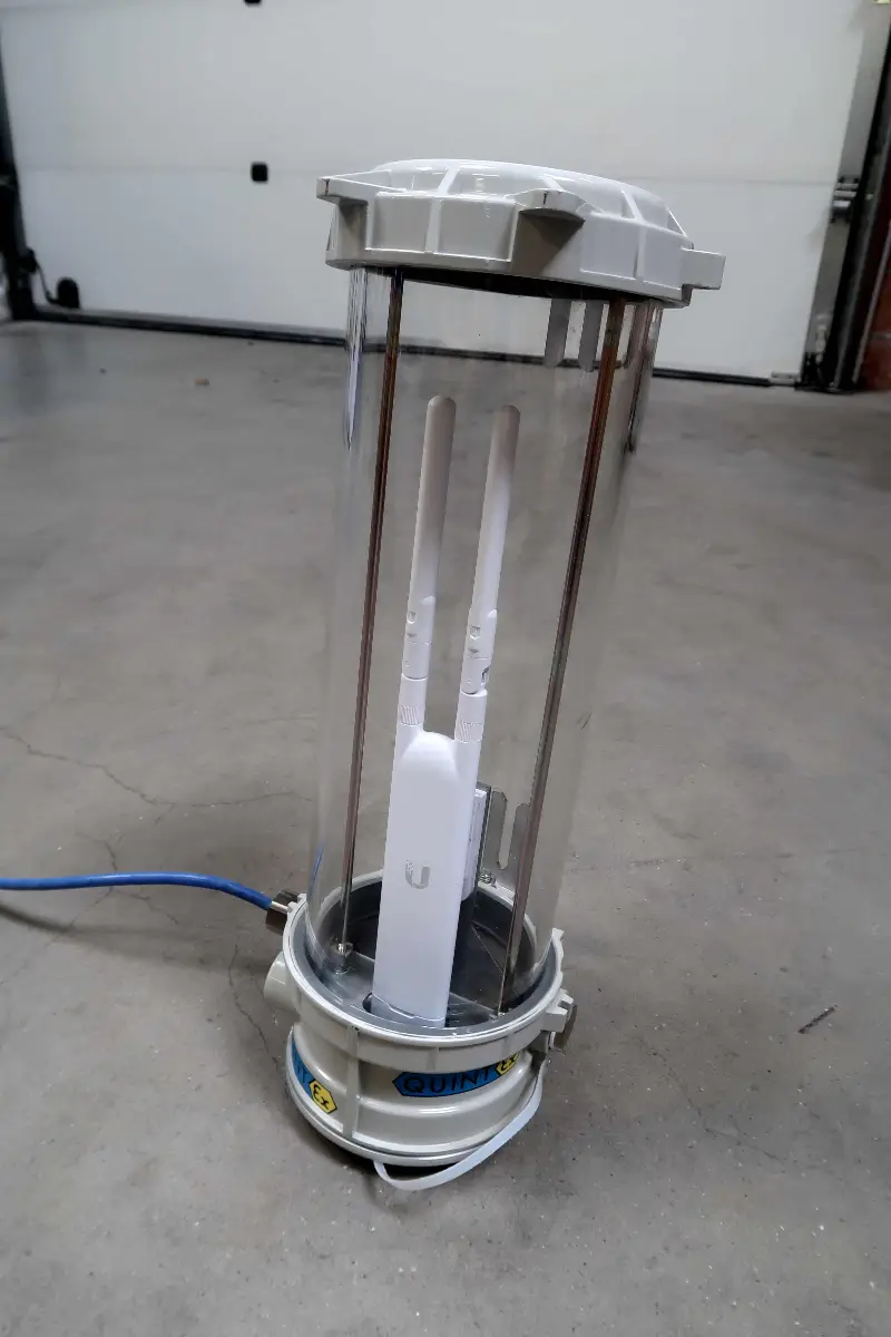 Een draadloze router zit in een doorzichtige cilindrische behuizing waaraan een kabel is bevestigd, en staat op een betonnen vloer voor een garagedeur.