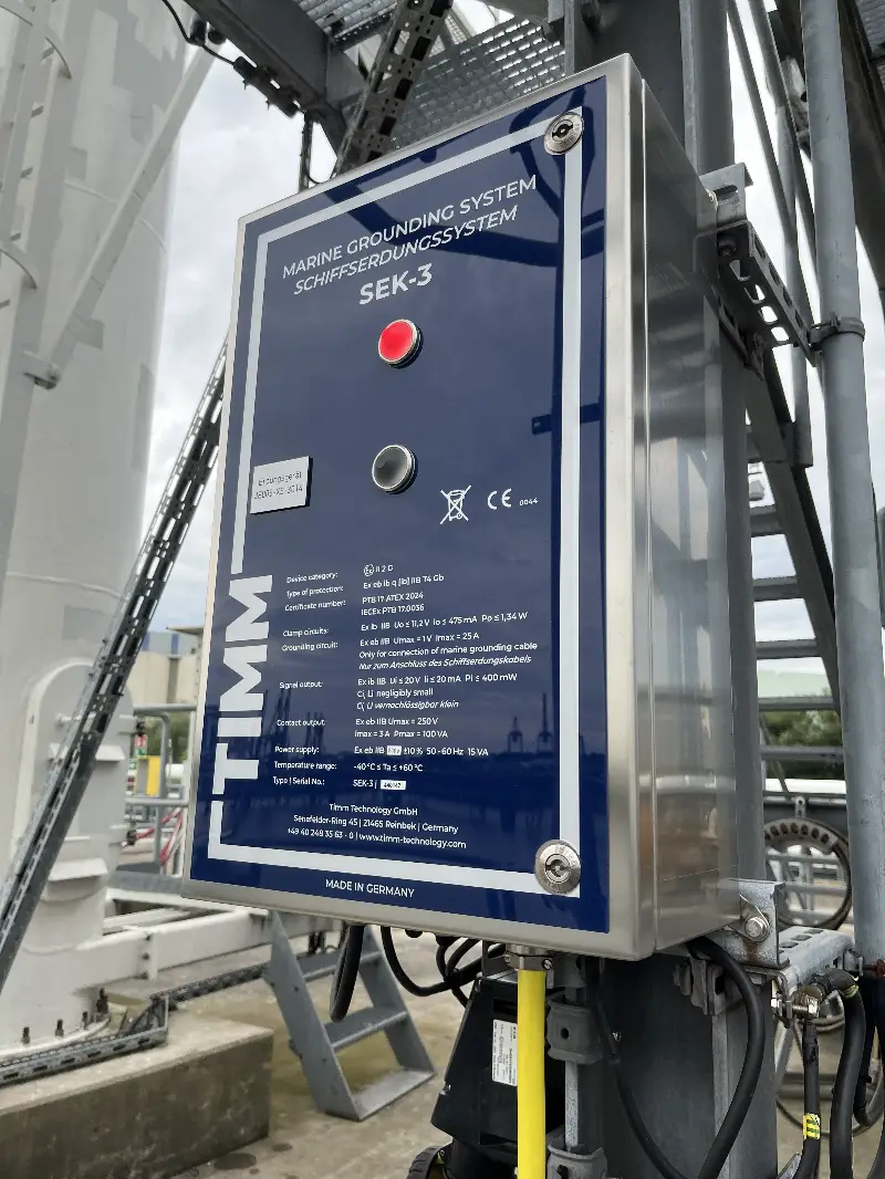 Een close-up van een blauwe schakelkast voor een maritiem aardingssysteem, met het opschrift „SEK-3“ van TIMM, die buiten op een metalen frame is gemonteerd, met tekst in het Duits en Engels, diverse technische details en zichtbare aangesloten kabels.