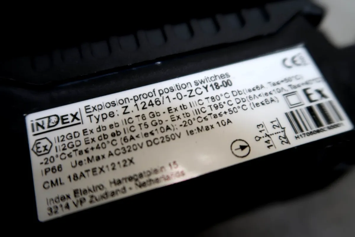 Close-up van een wit etiket op een zwart apparaat met daarop de technische specificaties van een explosieveilige positieschakelaar, waaronder gegevens over spanning, stroomsterkte, temperatuur en certificering. Er zijn ook wat tekst en logo’s te zien.