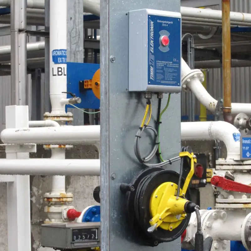 Op een metalen paal zijn een blauwe schakelkast en een gele oprolbare kabelhaspel bevestigd, omringd door industriële leidingen en kleppen die zijn voorzien van diverse labels, in een fabrieksomgeving.
