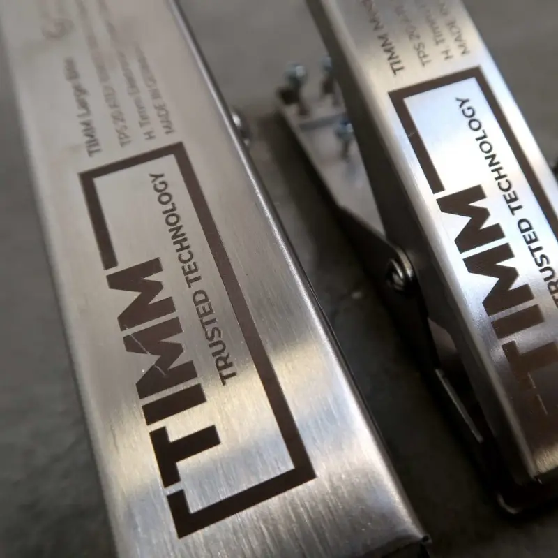 Close-up van twee voorwerpen van geborsteld metaal met het opschrift „TIMM TRUSTED TECHNOLOGY” en gegraveerde technische details, geplaatst op een grijs oppervlak.