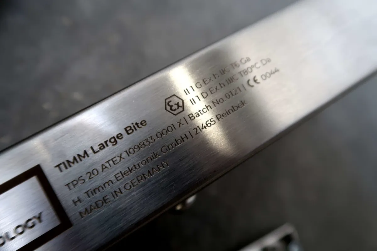 Close-up van een metalen apparaat met het opschrift „TIMM Large Bite“, waarop technische specificaties, certificeringssymbolen en productiegegevens, waaronder „Made in Germany“, in het oppervlak zijn gegraveerd.
