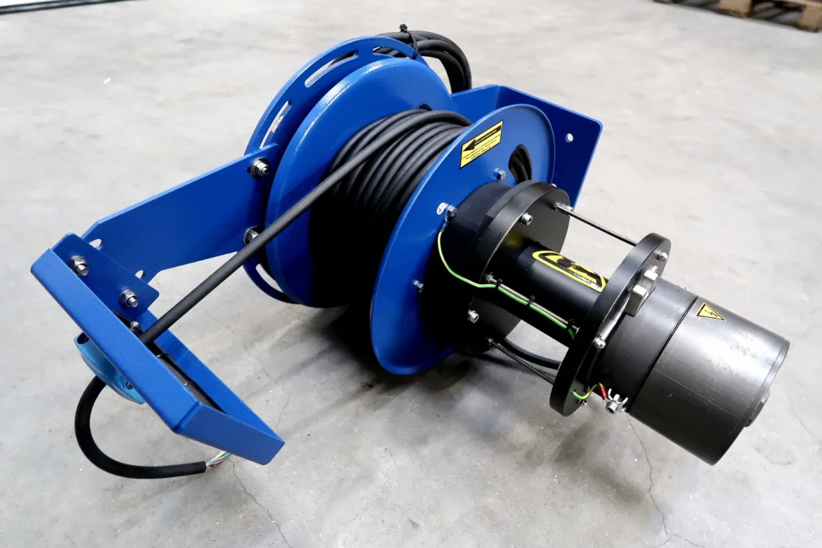 Een blauwe industriële kabelhaspel met een zwarte elektromotor eraan bevestigd, die op een grijze betonnen vloer ligt. De haspel is opgerold met zwarte kabel en is voorzien van waarschuwingslabels.