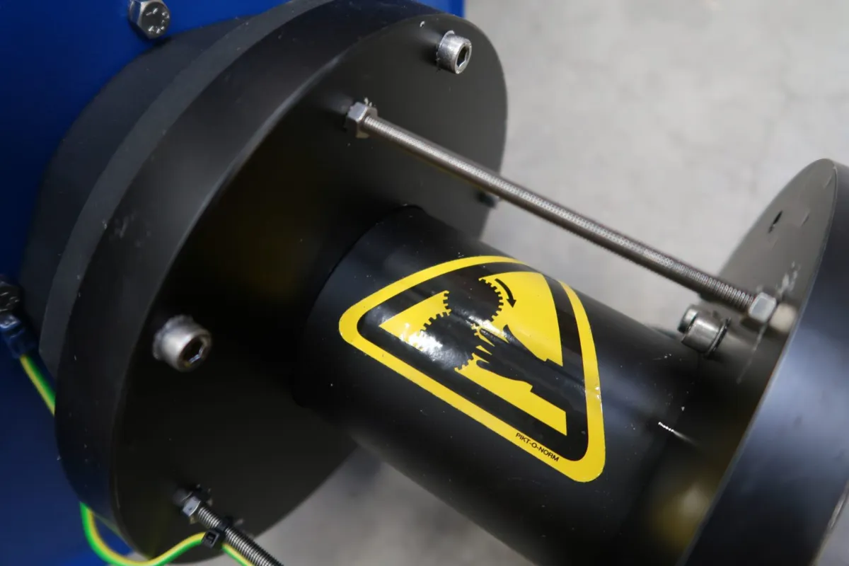 Close-up van een mechanisch onderdeel met een gele waarschuwingssticker waarop te zien is hoe een hand door tandwielen wordt bekneld, als waarschuwing voor het gevaar van beknellingspunten. Het onderdeel wordt bijeengehouden door bouten en is omgeven door metalen platen.