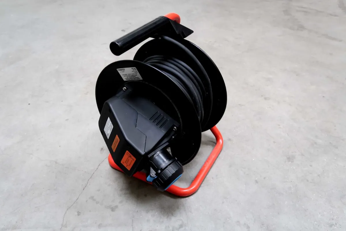 Op een grijze betonnen vloer staat een zwarte verlengsnoerhaspel met een handvat en stopcontacten. De haspel is op een rode metalen standaard bevestigd.