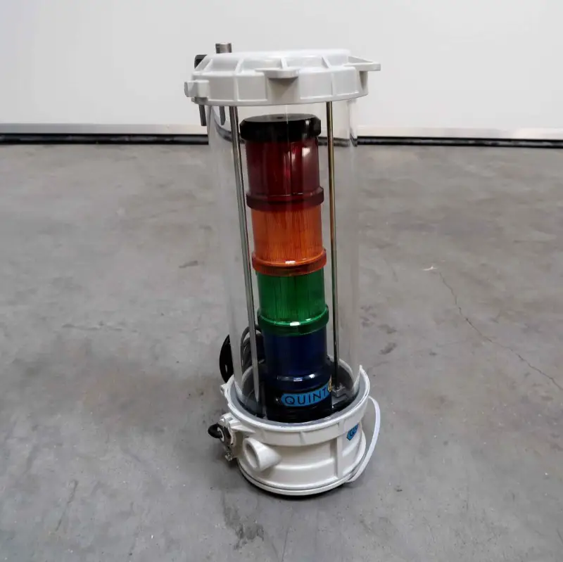 Een cilindervormig maritiem instrument met doorzichtige zijkanten, bestaande uit gestapelde rode, oranje en groene delen, staat op een grijze betonnen vloer tegen een effen muur.