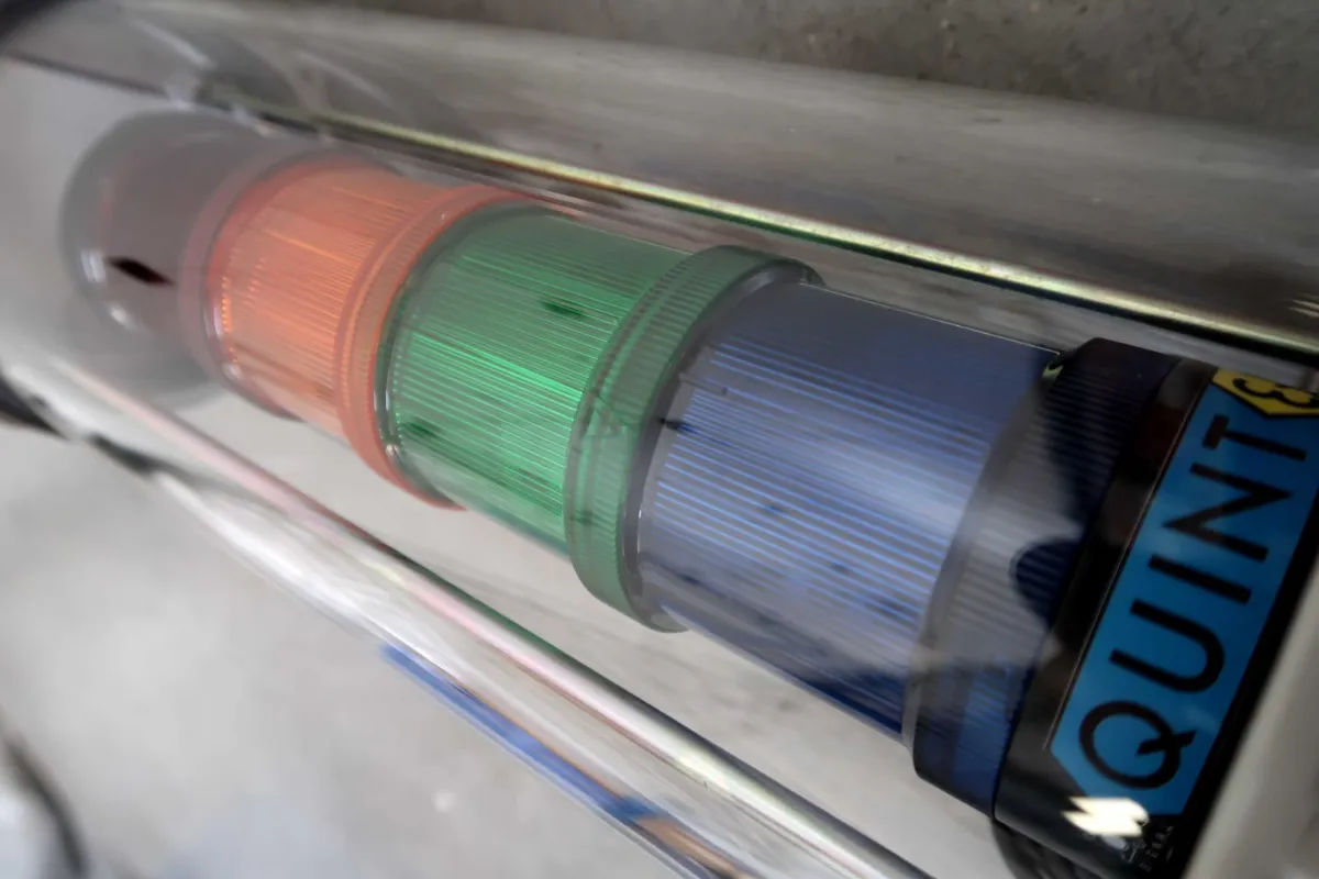Een close-up van een cilindervormig industrieel signaallicht met rode, groene en blauwe verlichte segmenten, voorzien van het opschrift QUINT, omhuld door een doorzichtige beschermkap.