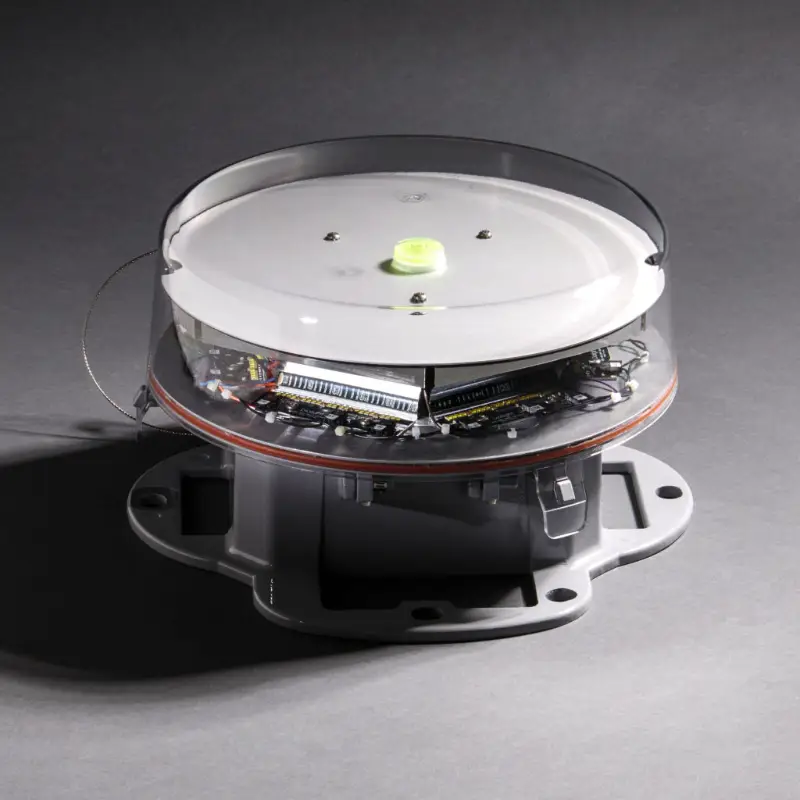 Een rond elektronisch apparaat met een doorzichtige bovenkant waardoor de interne onderdelen zichtbaar zijn, gemonteerd op een grijze voet, geplaatst op een grijs oppervlak met een schaduw aan de linkerkant.