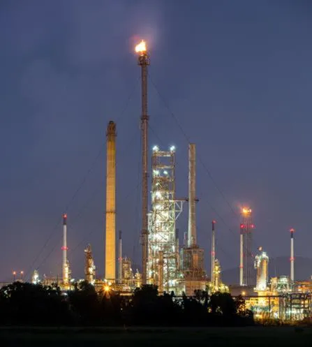 Een olieraffinaderij ’s nachts, met hoge torens en pijpleidingen die door lampen worden verlicht; op één toren wordt gas afgefakkeld, met een zichtbare vlam bovenaan tegen een donkere hemel.