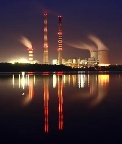 Een elektriciteitscentrale ’s nachts met drie schoorstenen waaruit rook opstijgt, verlicht door rode lampen, waarvan de weerspiegelingen zichtbaar zijn op het kalme water op de voorgrond.