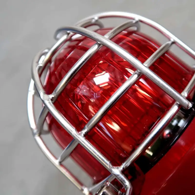Close-up van een rood noodwaarschuwingslicht met een metalen beschermkooi, waarop reflecties op het glasoppervlak te zien zijn.
