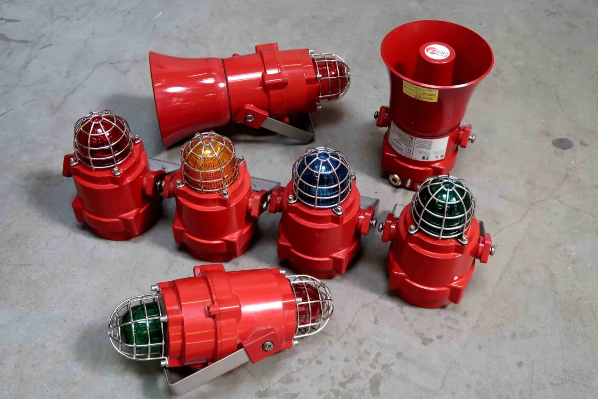 Op een grijze betonnen vloer staan zeven rode industriële waarschuwingslampen met metalen kooien, elk met een koepel in een andere kleur, en één rode sirene met een toeter.