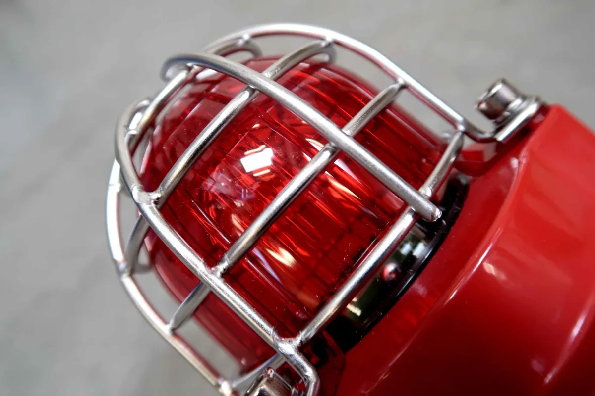 Close-up van een rood noodwaarschuwingslampje met een beschermende metalen kooi, afgebeeld tegen een wazige grijze achtergrond.