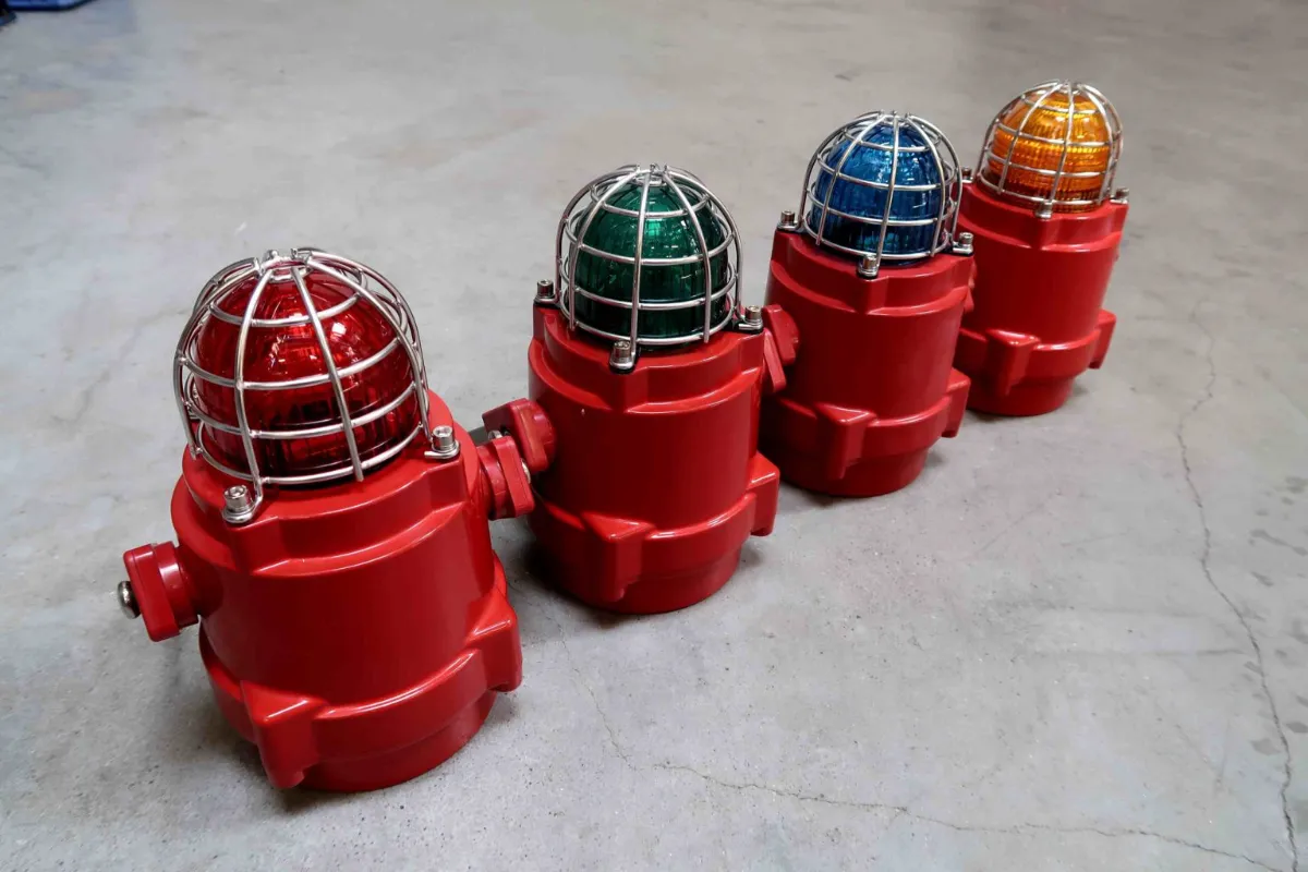 Vier industriële waarschuwingslampen met rode voetstukken en gekleurde koepels (rood, groen, blauw en oranje), afgeschermd door metalen kooien, die in een rij op een betonnen vloer staan opgesteld.