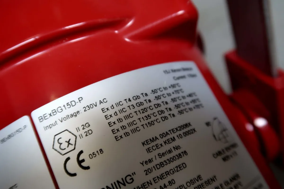 Close-up van een wit typeplaatje op een rood apparaat, waarop technische gegevens, certificeringssymbolen, informatie over de spanning en diverse veiligheids- en conformiteitsmarkeringen te zien zijn.