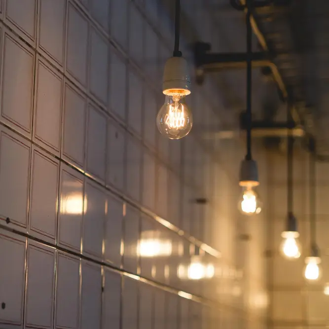 Langs een wit betegelde muur hangt een rij blootliggende gloeilampen aan het plafond, die een warme gloed verspreiden in een interieur in industriële stijl.