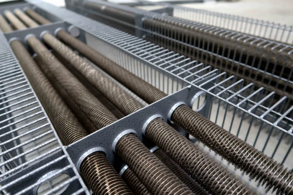 Close-up van industriële metalen buizen met opgerolde lamellen die door een metalen roosterframe lopen, waarschijnlijk onderdeel van een warmtewisselaar of een HVAC-systeem, in een fabriek of een technische omgeving.