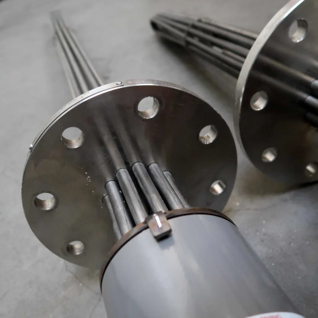 Close-up van twee industriële metalen verwarmingselementen met flenzen, meerdere staven en boutgaten, die op een grijs oppervlak liggen.