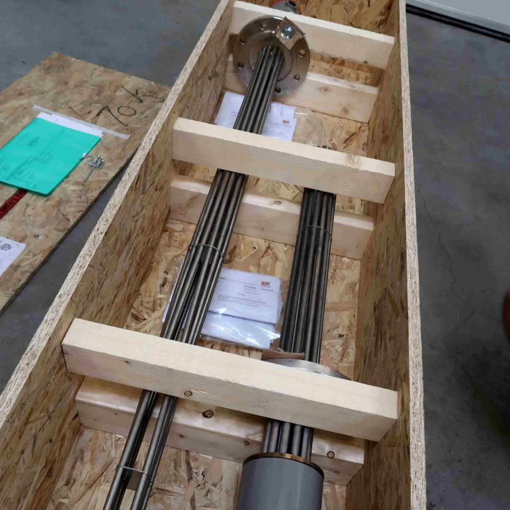 Metalen staven die in een houten kist zijn vastgezet, met daarnaast documenten en verpakkingsmateriaal, van bovenaf gezien op een betonnen vloer.
