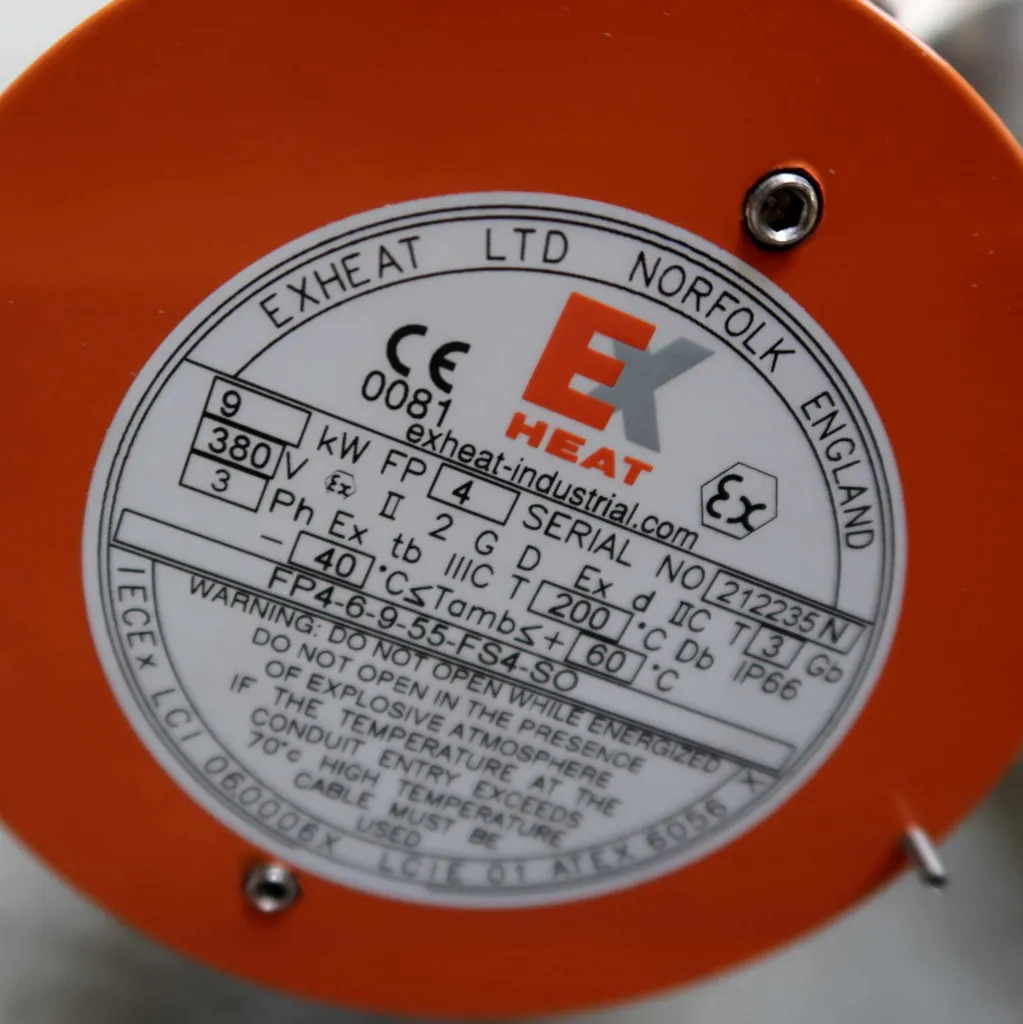 Close-up van een apparaatlabel met daarop de fabrikant EXHEAT LTD NORFOLK ENGLAND, modelgegevens, elektrische specificaties, certificeringen en de website. Het label is omrand door een oranje kader.
