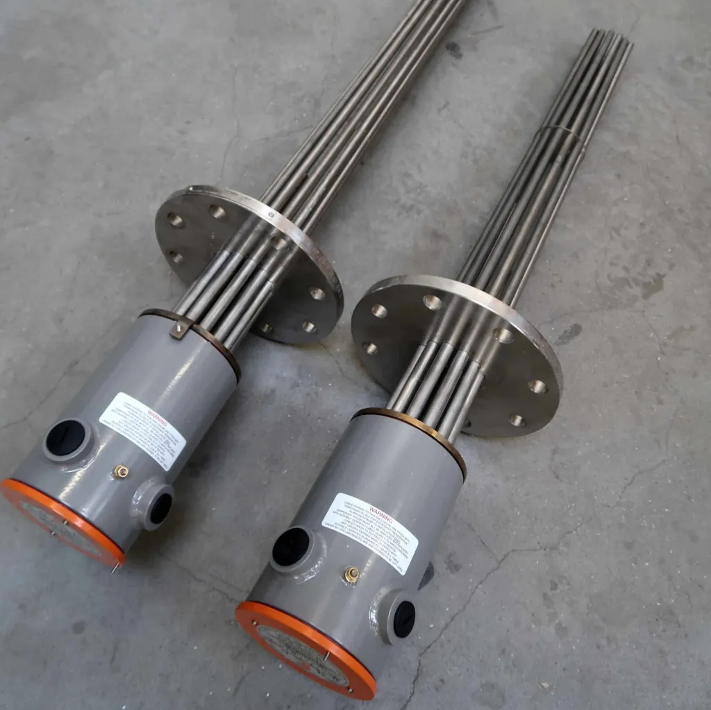 Op een grijze betonnen vloer liggen twee industriële buisvormige dompelverwarmers met metalen buizen en flensvoeten. Op de voeten staan labels en de cilindrische koppen zijn afgesloten met oranje eindstukken.
