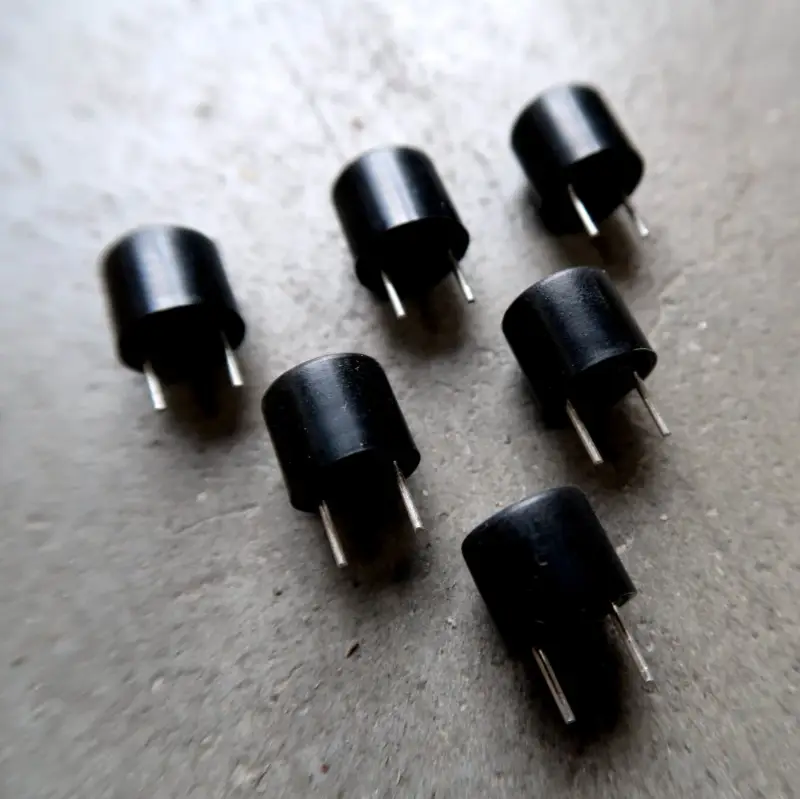 Zes kleine zwarte elektronische onderdelen met elk twee metalen pinnen zijn op een grijs oppervlak geplaatst en worden vanuit een hoek bekeken.