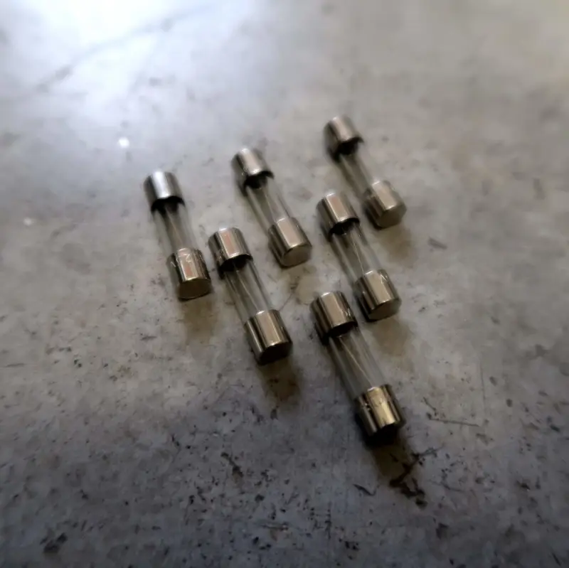 Zeven kleine, cilindervormige glazen zekeringen met metalen eindkappen staan op een grijs, licht gestructureerd oppervlak. De zekeringen liggen verspreid in een losse groep.
