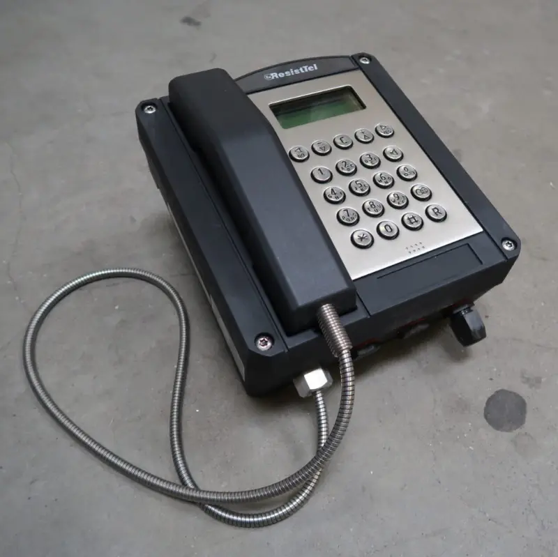 Op een betonnen vloer staat een robuuste telefoon in industriële stijl met een metalen toetsenbord en display. De hoorn is verbonden via een flexibel metalen snoer.