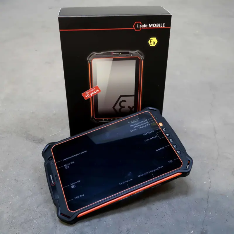 Op een grijs oppervlak ligt een robuuste tablet met oranje accenten voor zijn zwarte i.safe MOBILE-doos. Het scherm van de tablet is donker en weerkaatst het licht, en op de doos staan een afbeelding van het apparaat en de Ex-veiligheidsmarkering.