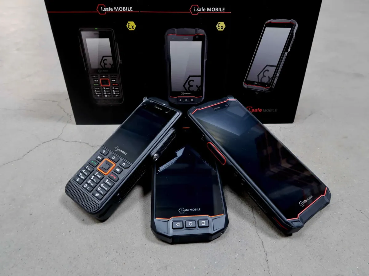 Op een betonnen ondergrond staan drie robuuste mobiele telefoons uitgestald, met op de achtergrond twee rechtopstaande productdozen, allemaal voorzien van het label i.safe MOBILE. De telefoons hebben een robuust ontwerp in zwart en oranje.