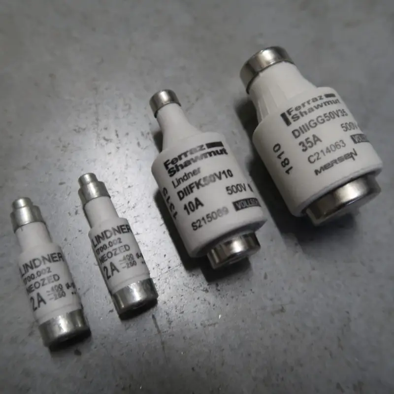 Op een grijs oppervlak liggen vier cilindervormige elektrische zekeringen van verschillende afmetingen. Elke zekering heeft een witte behuizing met metalen uiteinden en opgedrukte labels waarop de stroomsterkte staat aangegeven, zoals 2 A, 10 A en 35 A.