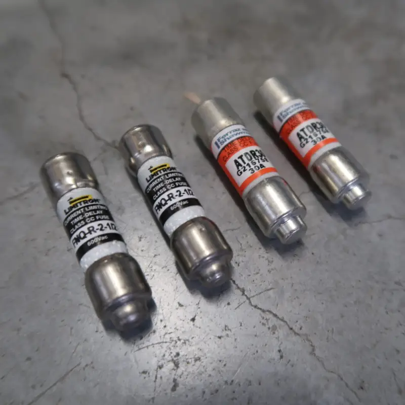 Vier cilindervormige zekeringen met metalen uiteinden en gelabelde behuizingen – twee met zwarte labels en twee met oranje labels – liggen op een grijs, gebarsten betonnen oppervlak.