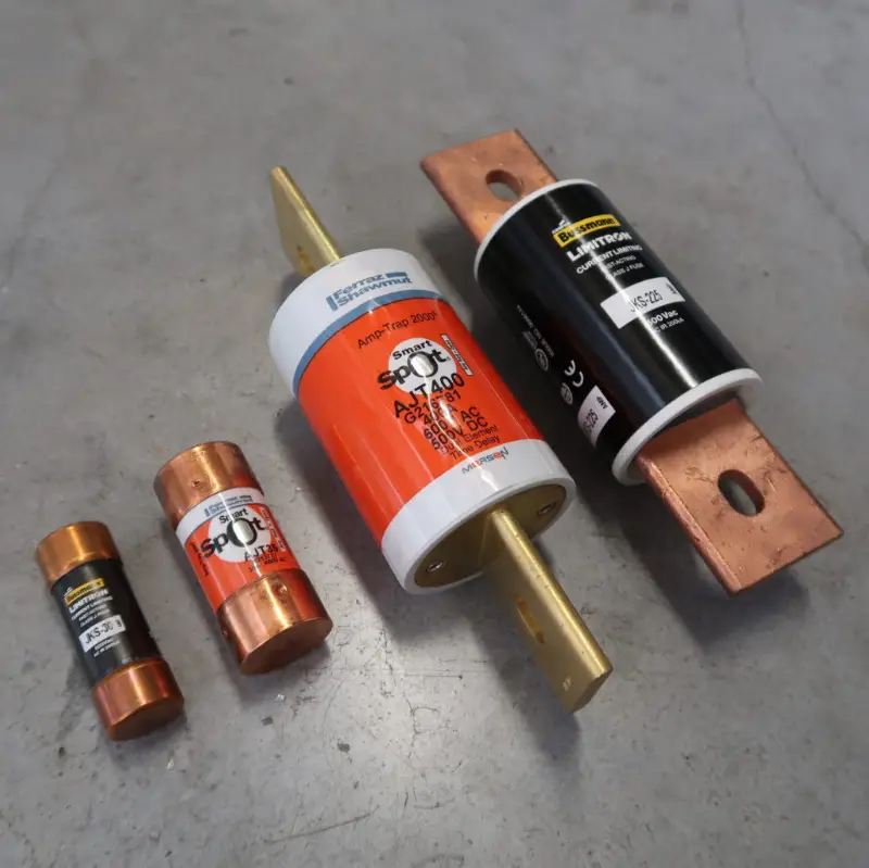 Vier elektrische zekeringen van verschillende afmetingen, waarvan twee met koperen uiteinden en twee met grote metalen lamellen, liggen op een grijs oppervlak. De zekeringen zijn voorzien van specificaties en hebben oranje, witte en zwarte omhulsels.
