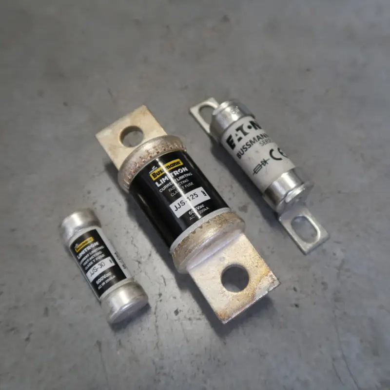 Op een grijs oppervlak liggen drie industriële zekeringen van verschillende afmetingen. Twee daarvan zijn klein en hebben metalen uiteinden, en één is groter, met een zwart-wit etiket en platte bevestigingsbeugels.
