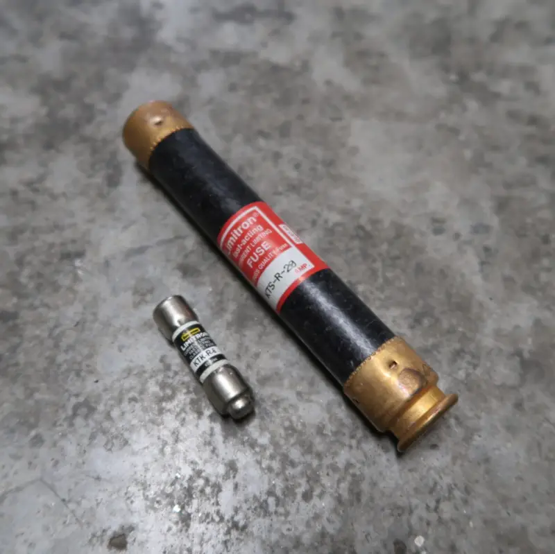Op een grijs, gestructureerd oppervlak liggen twee cilindervormige zekeringen. De grotere zekering is zwart met messing uiteinden en een rood etiket, terwijl de kleinere zekering metaalachtig is met zwarte markeringen.