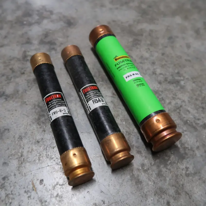 Er liggen drie cilindervormige elektrische zekeringen met metalen uiteinden op een grijs oppervlak. Twee zijn zwart met witte labels, en één is groen met een geel label.