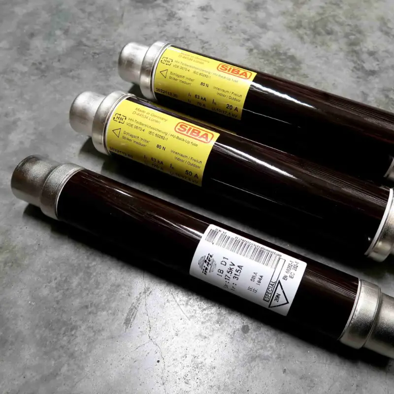Op een grijs oppervlak liggen drie cilindervormige elektrische zekeringen met gele SIBA-etiketten en metalen dopjes. De zekeringen liggen diagonaal ten opzichte van elkaar, waarbij één etiket gedeeltelijk zichtbaar is.