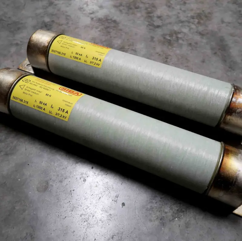 Twee cilindrische hoogspanningszekeringen met gele etiketten, metalen doppen aan beide uiteinden en grijze behuizingen liggen naast elkaar op een grijs oppervlak met structuur.