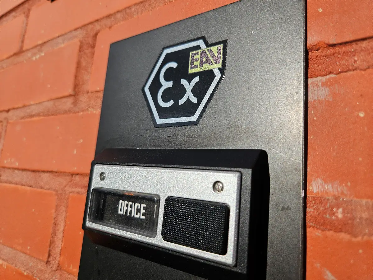 Op een bakstenen muur is een zwart kantoorbord met een luidspreker bevestigd. Daarboven bevindt zich een zeshoekig symbool met de letters ‘Ex’ en een klein ‘EN’-label. De foto is schuin vanuit een hoek genomen in fel zonlicht.