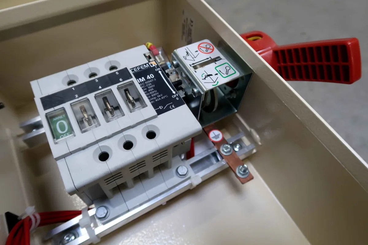 Close-up van een schakelkast met stroomonderbrekers, gelabelde schakelaars en een rode hendel voor het bedienen van de stroomonderbreker, gemonteerd in een metalen behuizing.