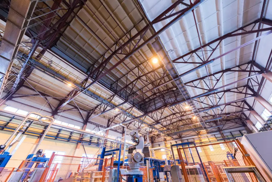 Het interieur van een grote industriële fabriek met stalen balken, machines en apparatuur onder een hoog, goed verlicht plafond.